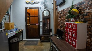 A compact room with a wooden door, a clock, a fan, a small Christmas tree, and a cabinet with numbered drawers. - Sip N' Camp - Hostel (Bangkok)