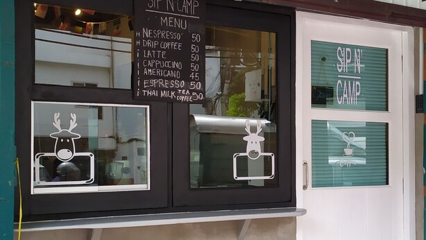 A coffee shop with a menu board listing items like Nespresso, drip coffee, and latte. There are decorative reindeer cutouts on the windows. - Sip N' Camp - Hostel (Bangkok)