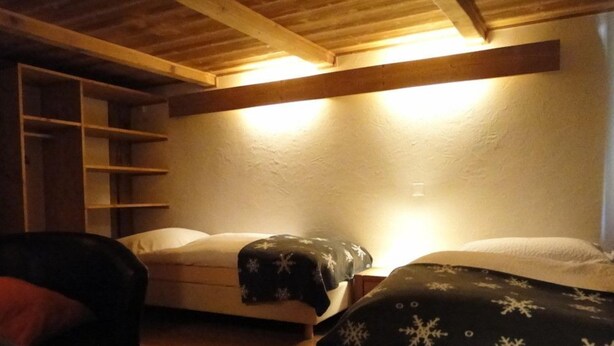 Een knus vertrek met houten balken, een bed met een deken met sneeuwvlokpatroon, een stoel en een wandplank.