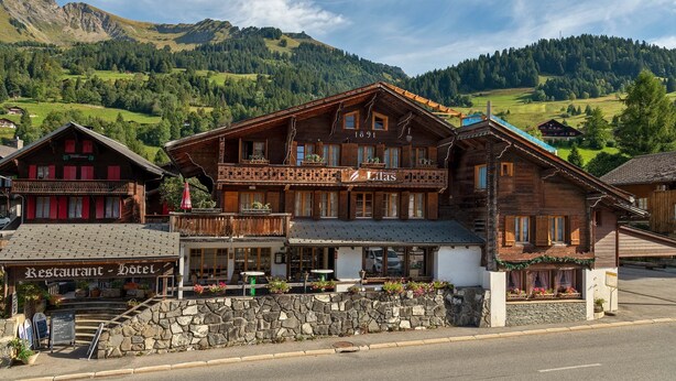 Un bâtiment traditionnel en bois, doté d’un balcon, d’une enseigne indiquant « Restaurant - Hôtel » et d’un mur de pierre à l’avant.