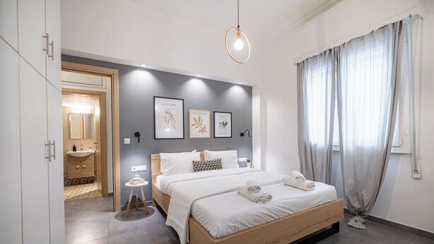 A modern bedroom with a wooden bed, a bedside table, a nightstand, a bathroom visible through an open door, and a large window with curtains. - Cosy apt near Piraeus central port, Mikrolimano port & Pasalimani port. (Pireas)