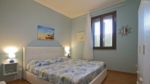 A bedroom with a bed, two bedside tables with lamps, a window with blinds, and a wall-mounted mirror.