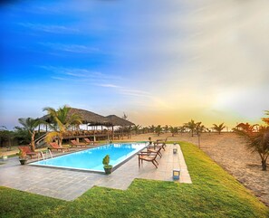 Un área de piscina con sillones de descanso y sombrillas, rodeada de palmeras y una playa.