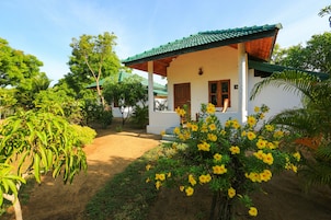 Una casita blanca con techo verde, rodeada de abundante vegetación y flores amarillas.