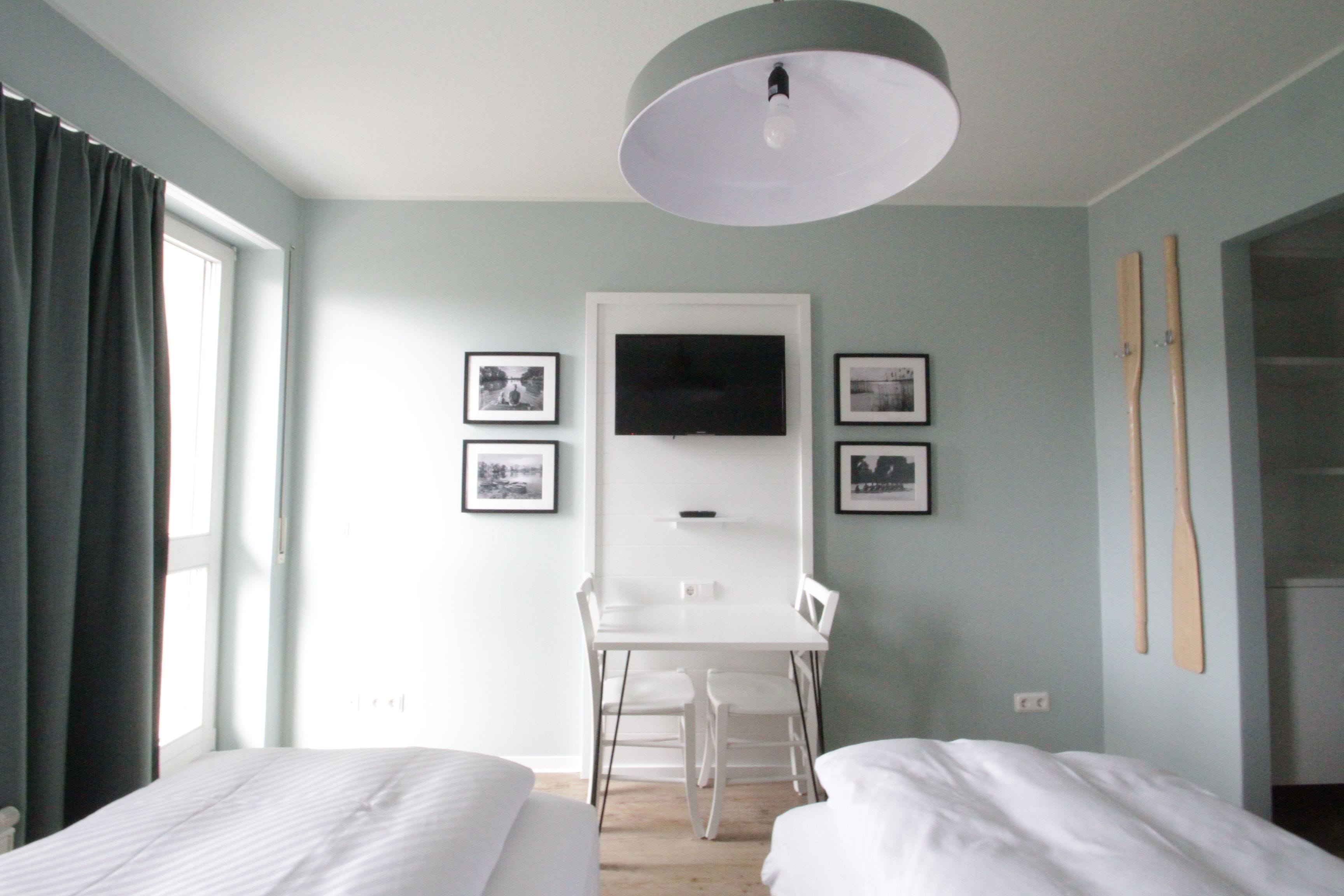 A hotel room with two beds, a desk, a TV, and framed pictures on the wall.