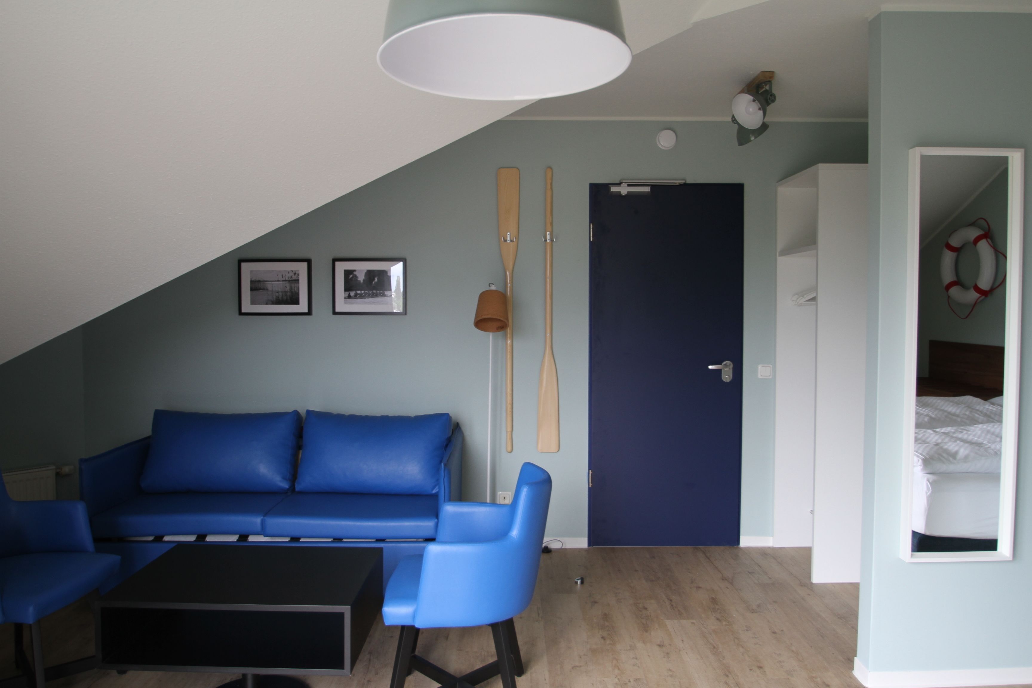 Ein Zimmer mit einem blauen Sofa, einem schwarzen Couchtisch, einer Tür mit einem Rettungsring und zwei Holzschlägern an der Wand.