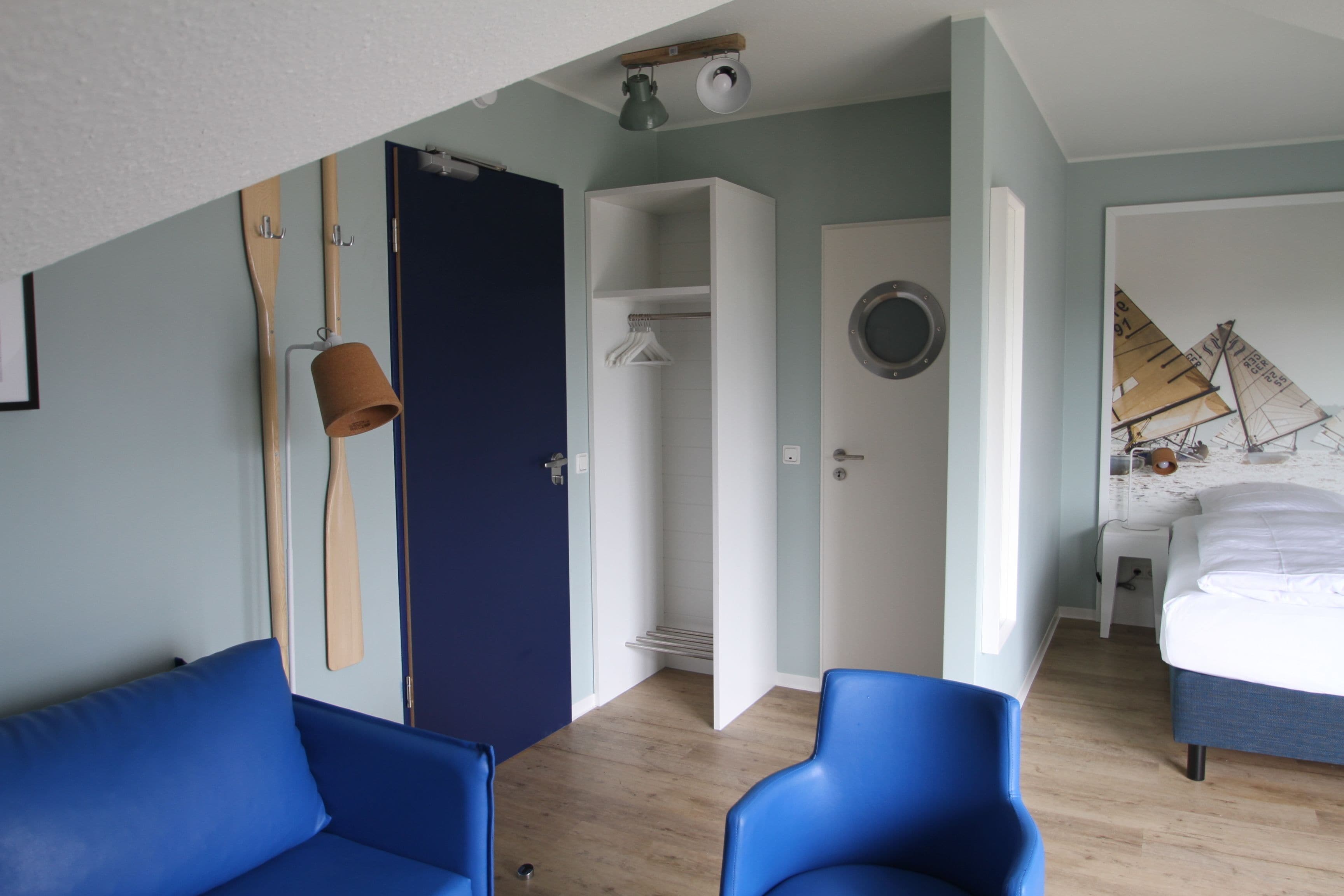 Una habitación con una cama, dos sillas azules, un armario, una puerta y una pared con una pintura de un velero.