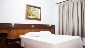 A neatly made bed with white linens, a wooden headboard, and a framed painting of flowers on the wall. - Js Palace Hotel (Alfenas)