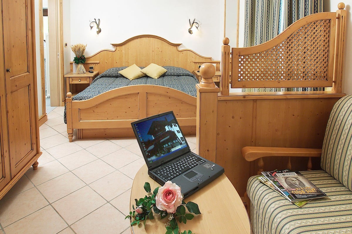 Quarto com cama de madeira, um laptop sobre uma mesinha, uma cadeira e uma janela com cortinas.