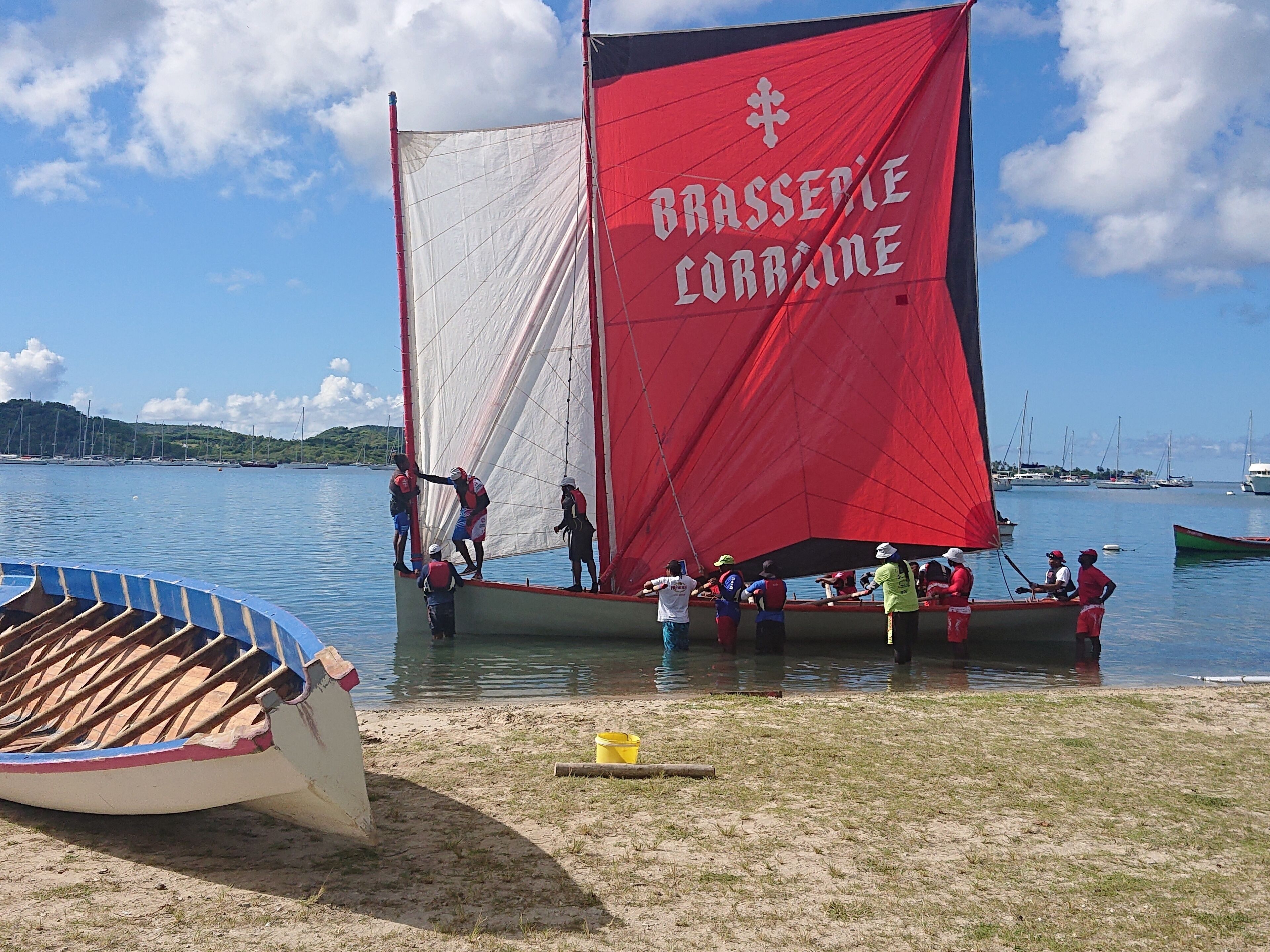 A sailboat with a red sail and the text "Brasserie Corbin" on it, docked at a beach with a small boat nearby.