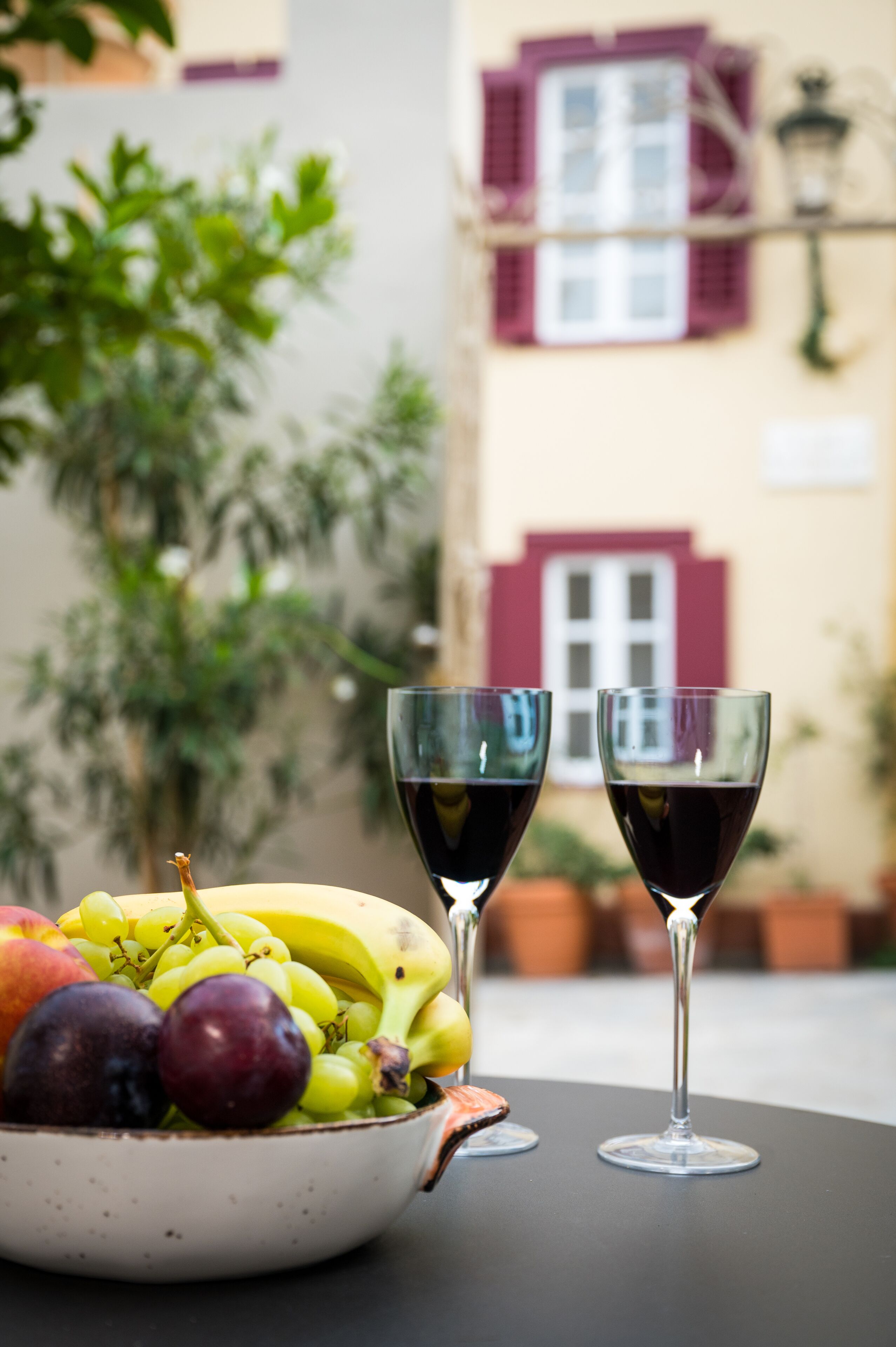A bowl of fruit and two wine glasses on a table with a building in the background.