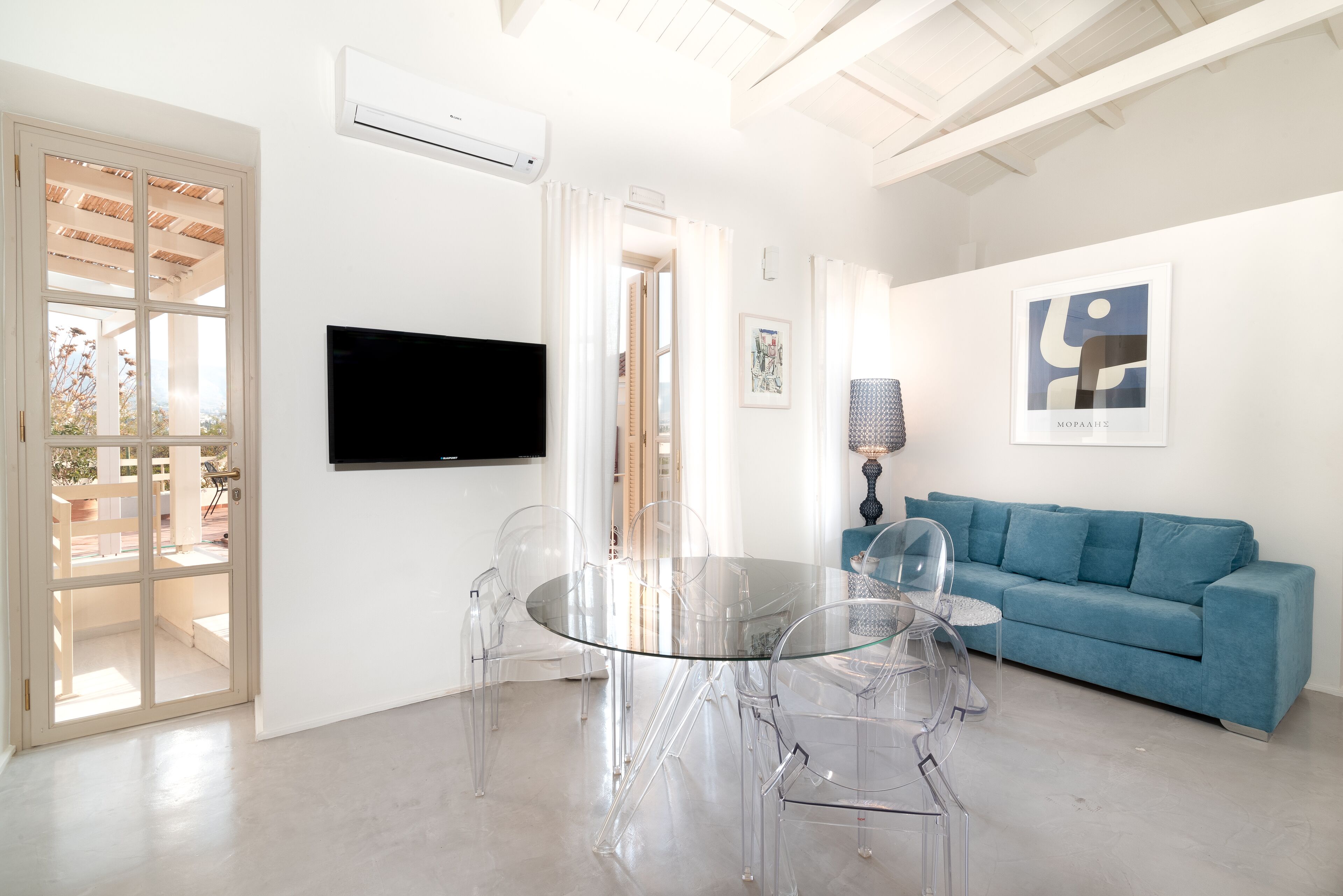 Un salon moderne avec une table à manger en verre, des chaises transparentes, un canapé bleu et un téléviseur fixé au mur.