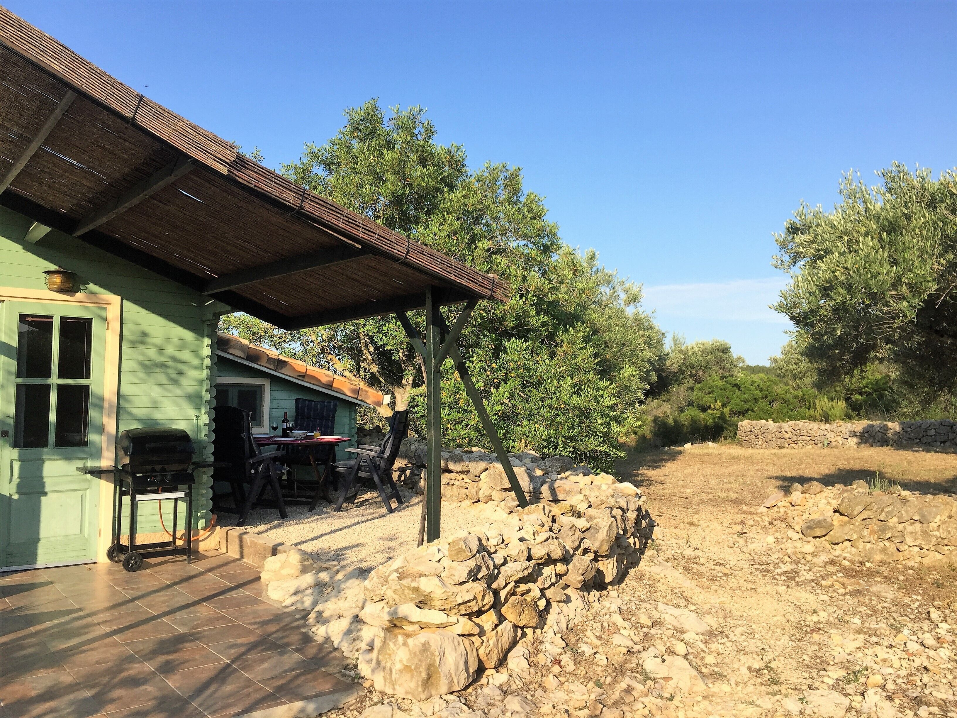 Un área de estar al aire libre rústica con una mesa, sillas, un muro de piedra y un edificio verde con suelo de baldosas.