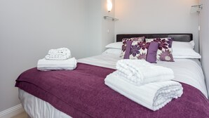 A neatly made bed with white and purple linens, a purple throw blanket, and a glass shelf with a white towel. - Angel Mews (Poole)