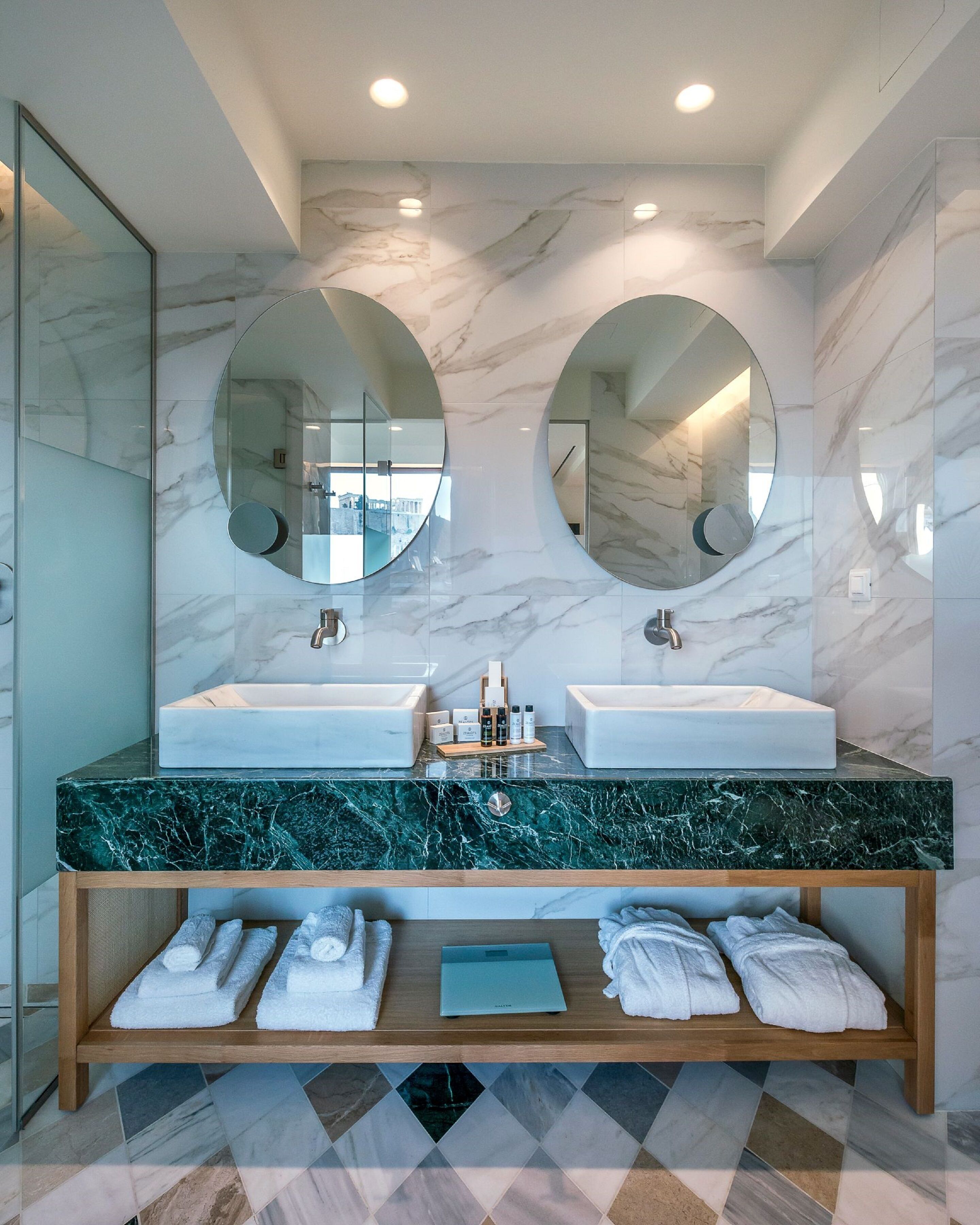 Un baño moderno con dos lavamanos, encimeras de mármol y espejos grandes. Hay toallas y artículos de aseo en el mueble.