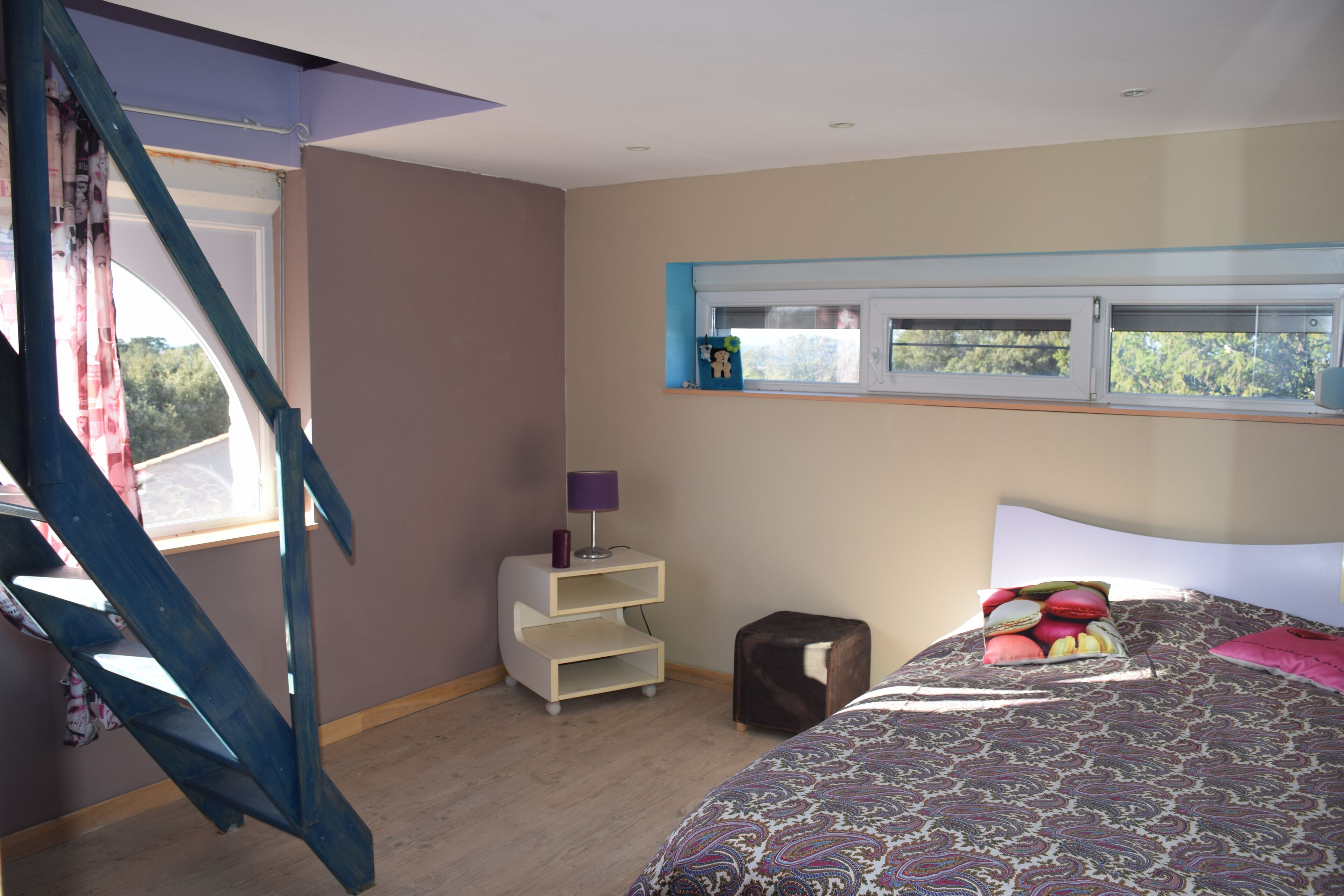 Une chambre à coucher avec un lit, une table de chevet, un petit tabouret, une fenêtre et un escalier.