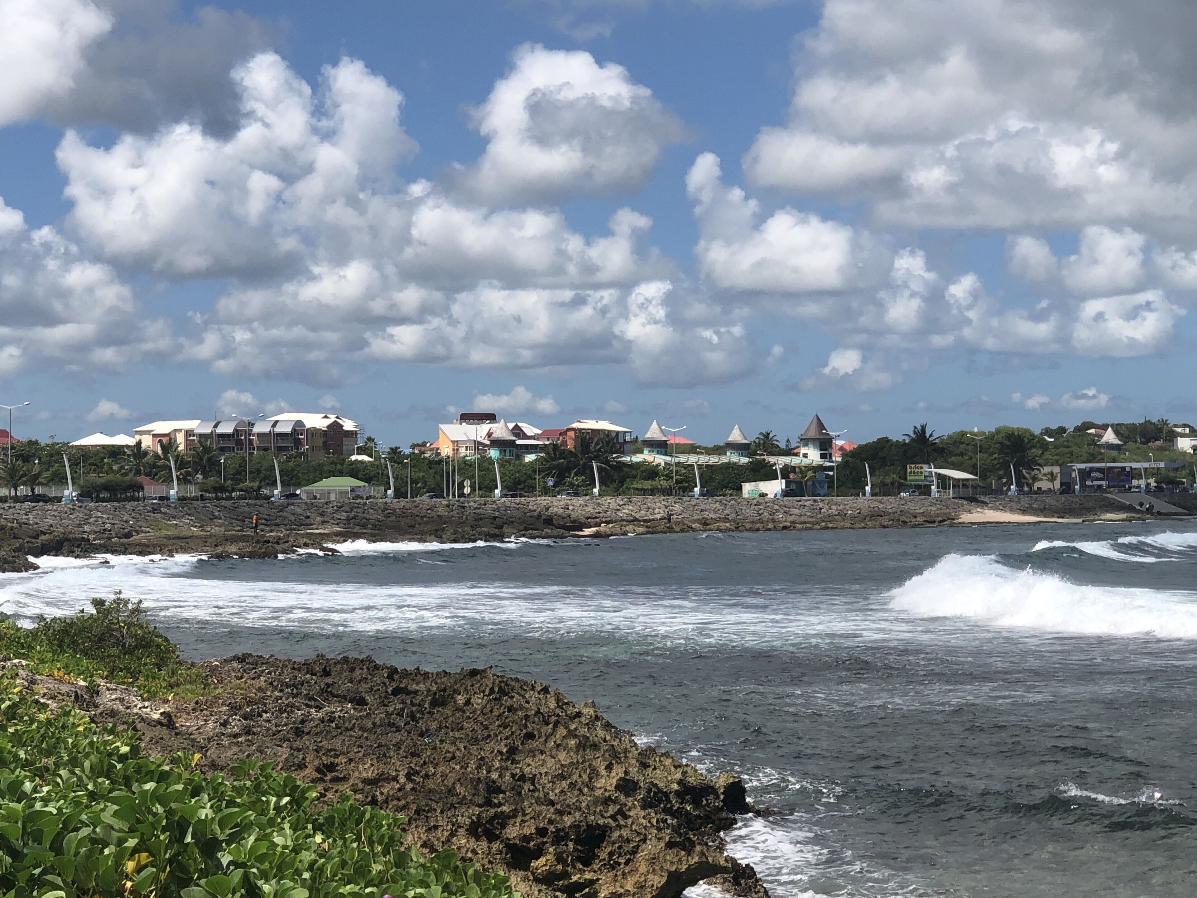 A coastal view with waves crashing against a rocky shore, buildings in the background, and a clear sky with clouds.