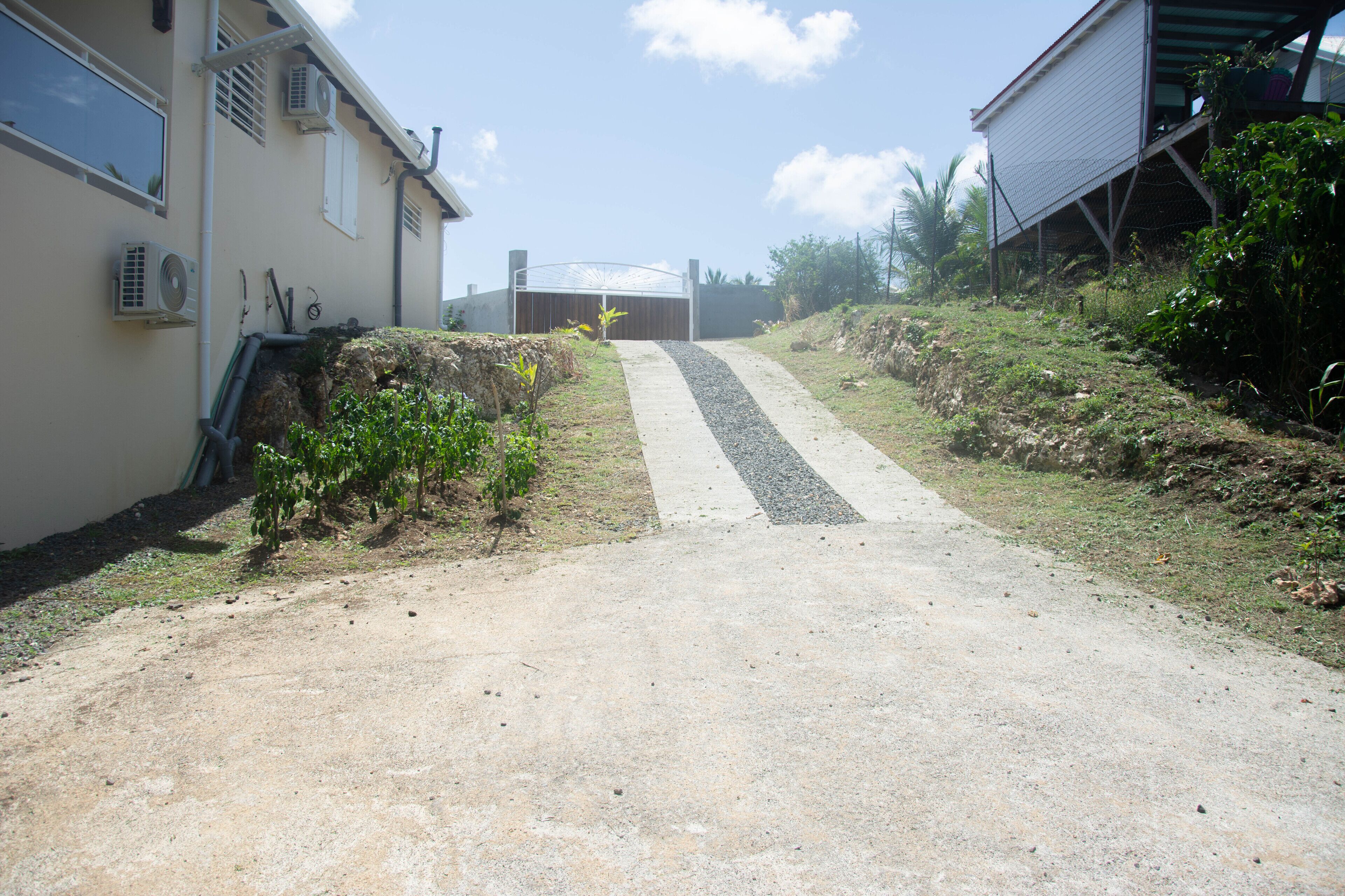 Un camino estrecho y sin asfaltar que conduce a una entrada porticada con techo metálico, rodeado de vegetación y un edificio con unidades de aire acondicionado.