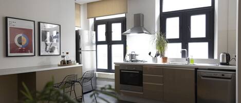 Eine moderne Küche mit Kochinsel, integriertem Backofen und Geschirrspüler. An der Wand befinden sich zwei gerahmte Bilder, ein Topf mit Pflanze und ein Fenster mit schwarzen Rahmen.