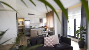 A modern living room with a dark sofa, a coffee table, and a dining area with a table and chairs. There is a kitchen in the background with a refrigerator and a window with blinds. - GALATA M84  (Istanbul)