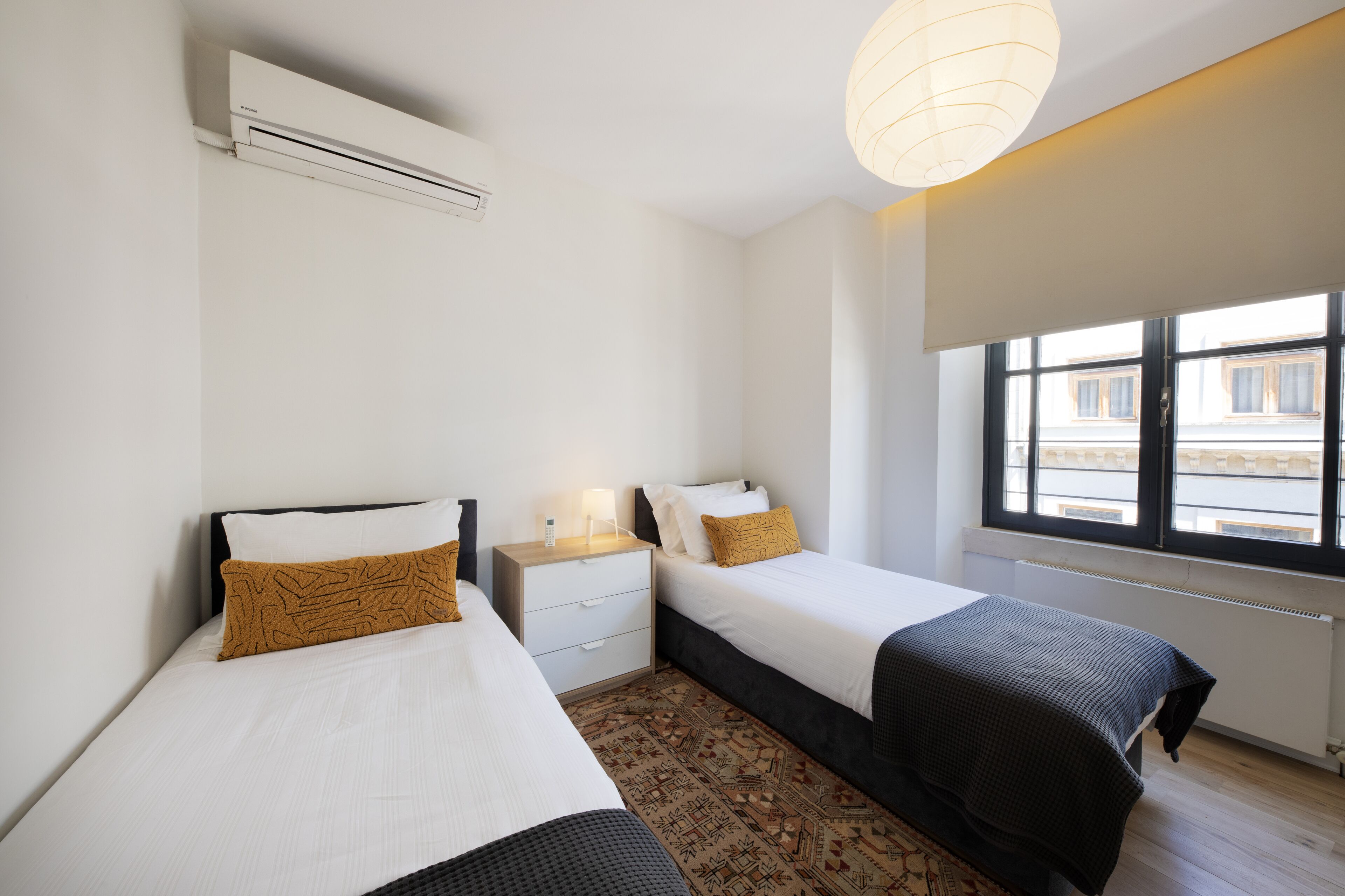 Quarto de hotel com duas camas, uma mesa de cabeceira, uma luminária e janela com vista para prédios.