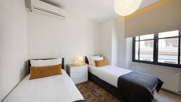 Quarto de hotel com duas camas, uma mesa de cabeceira, uma luminária e janela com vista para prédios.