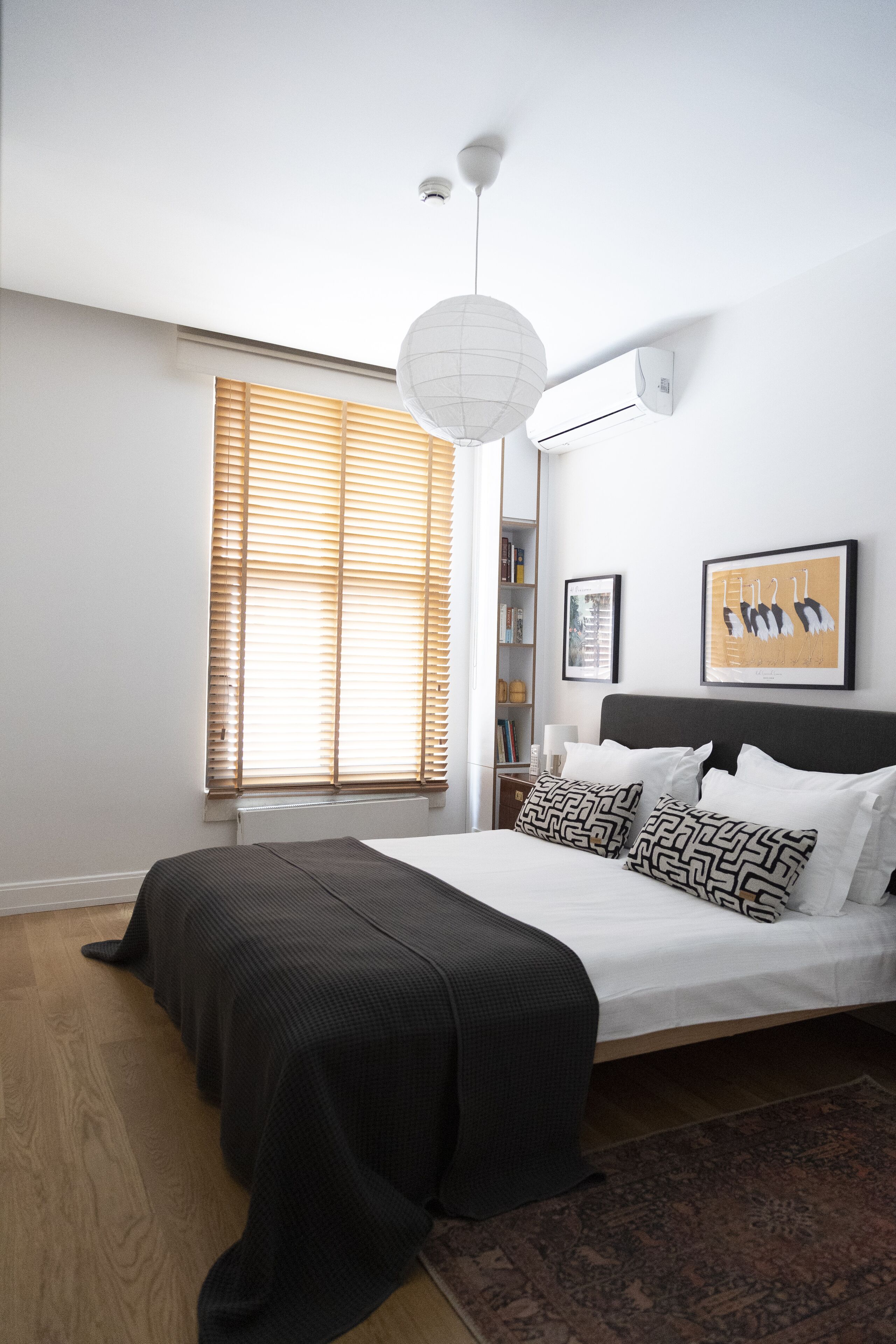 A bedroom with a large bed, a window with blinds, a ceiling light, and a wall-mounted air conditioner.