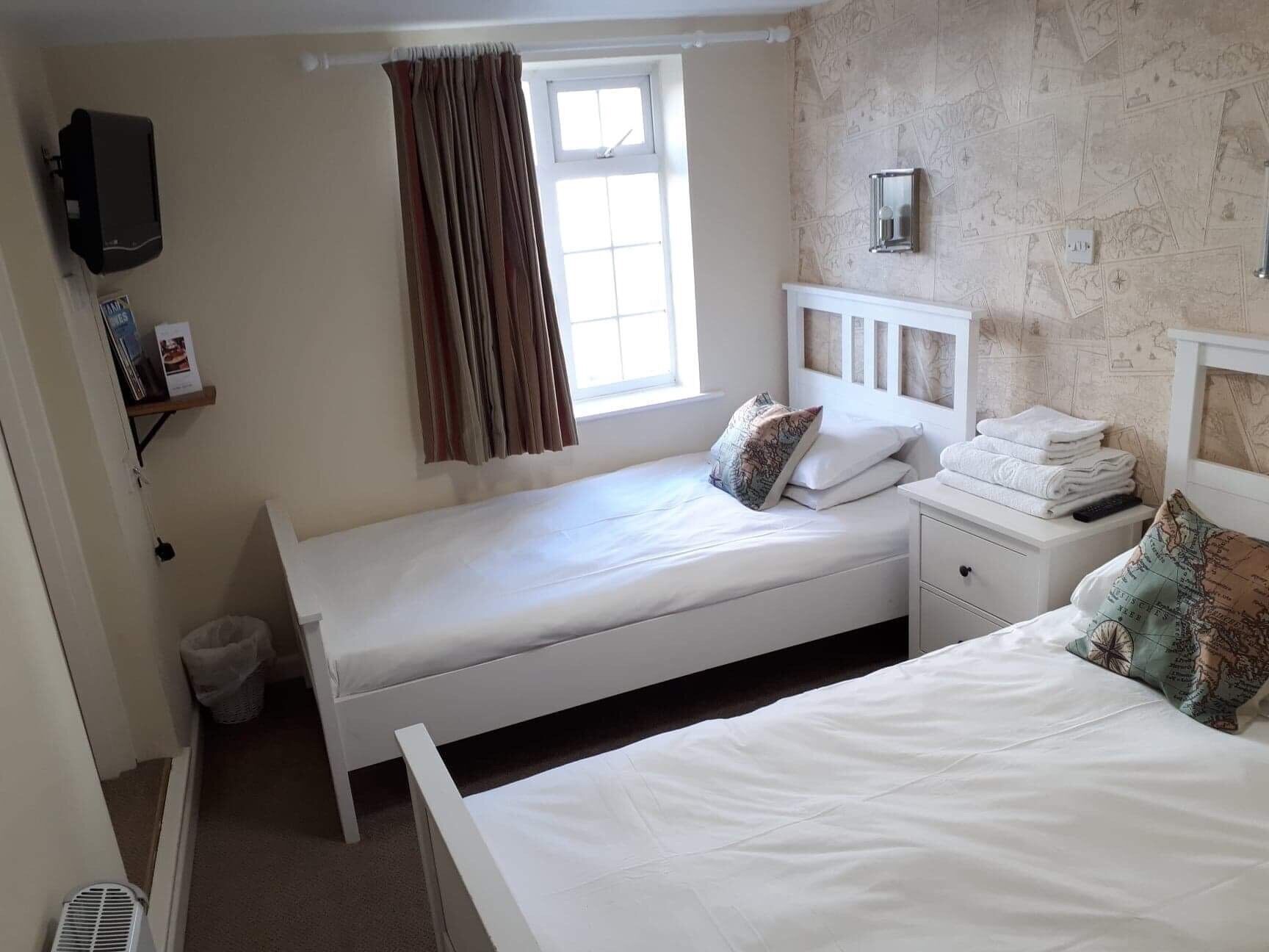 A small, clean room with two single beds, a window with curtains, a wall-mounted TV, and a small shelf with books.