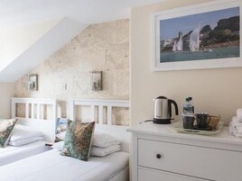 Quarto de hotel com duas camas, cômoda branca, pintura de uma paisagem costeira e vista para o mar.