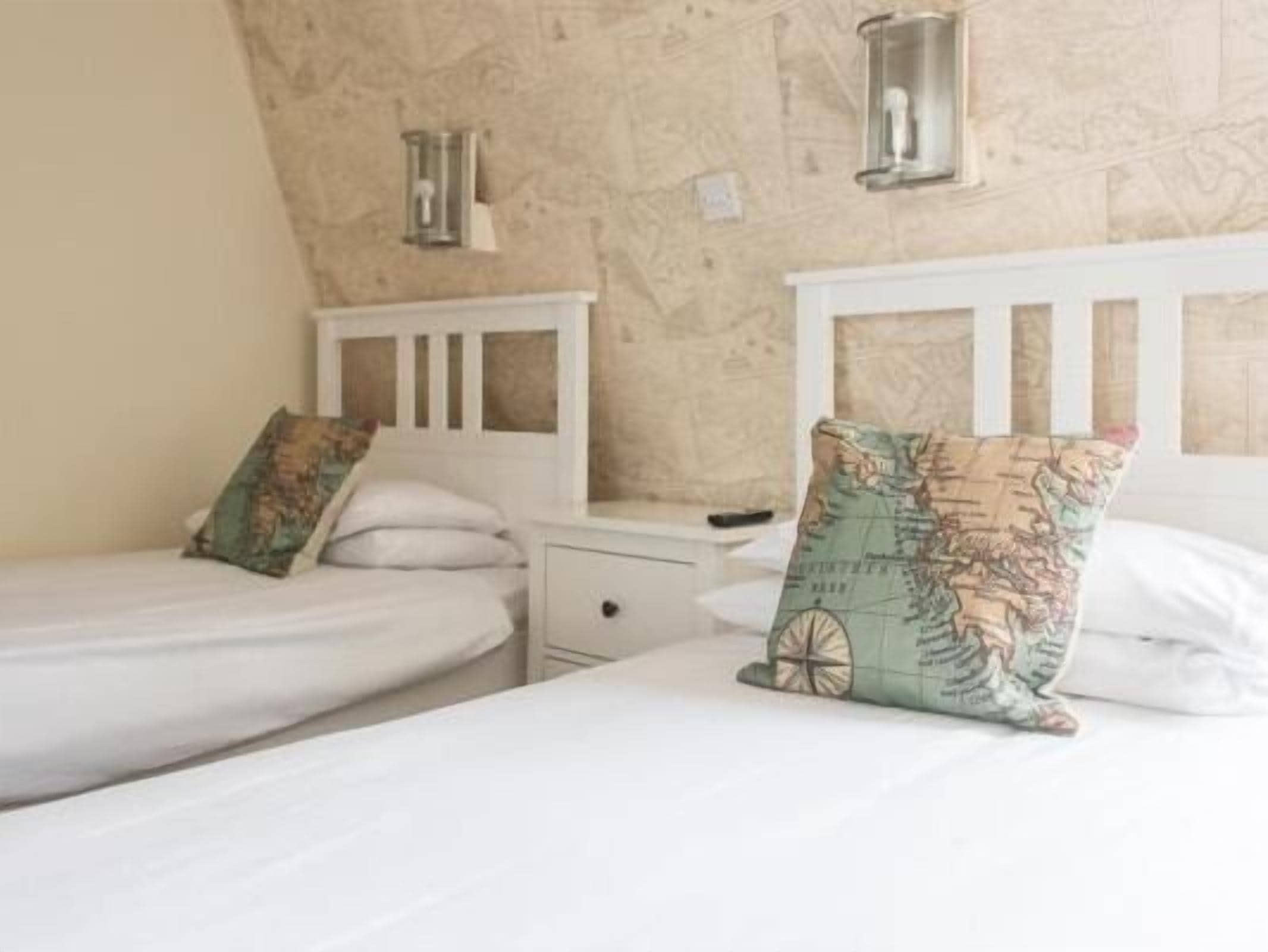 A bedroom with two single beds, each with a white headboard and a small bedside table. There are two wall-mounted lamps on the wall above the beds.