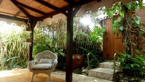 A covered patio with a wicker chair, surrounded by lush tropical plants and a wooden door.