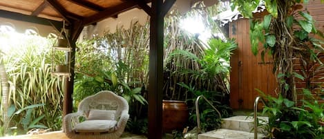 A covered patio with a wicker chair, surrounded by lush tropical plants and a wooden door.