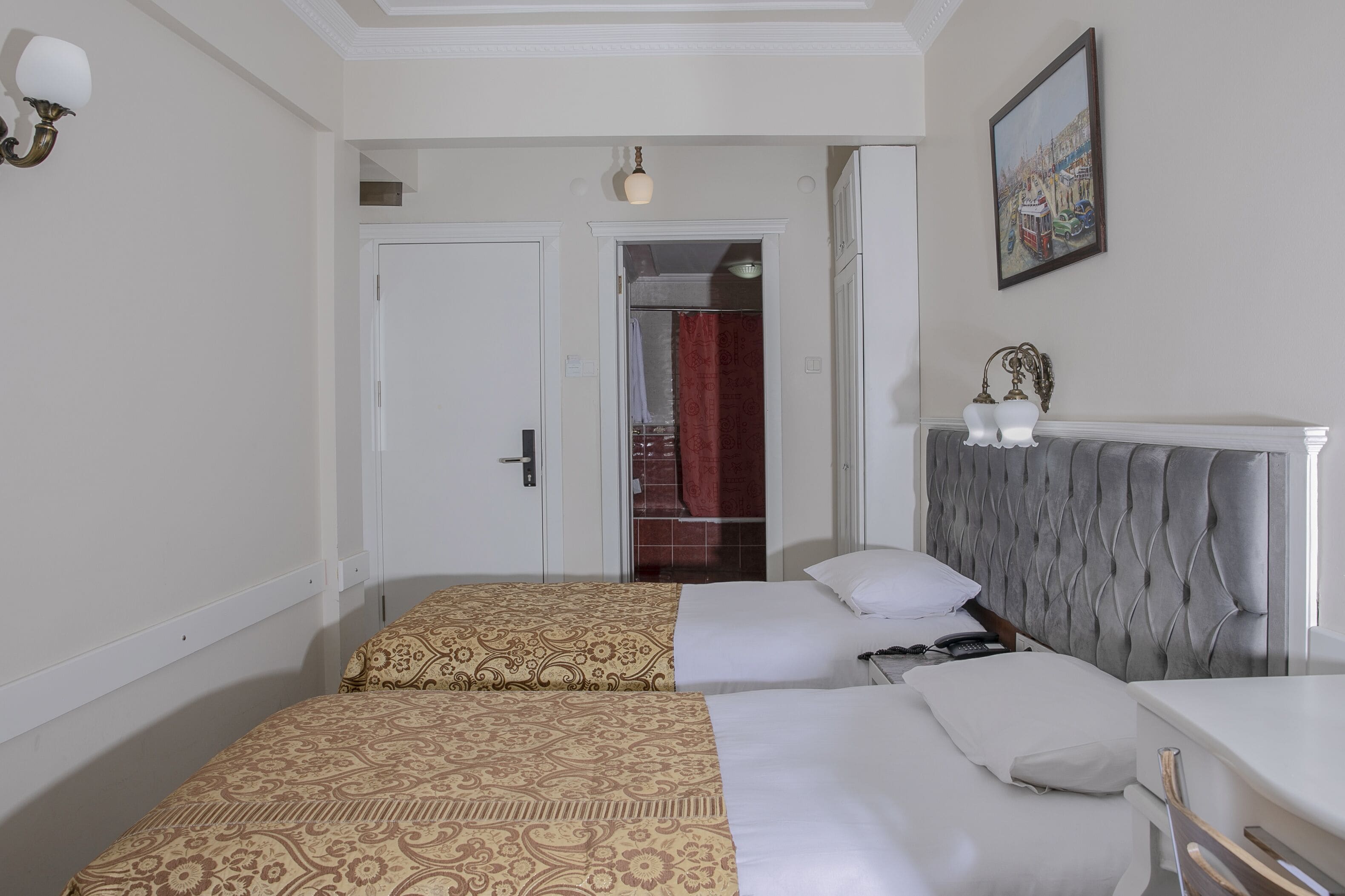 Habitación de hotel con dos camas, cabecero tapizado en gris, un cuadro en la pared y una puerta que da a un baño.