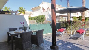Un patio exterior con mobiliario de mimbre, una mesa con tazas y una planta, y una pérgola de madera con una sombrilla.