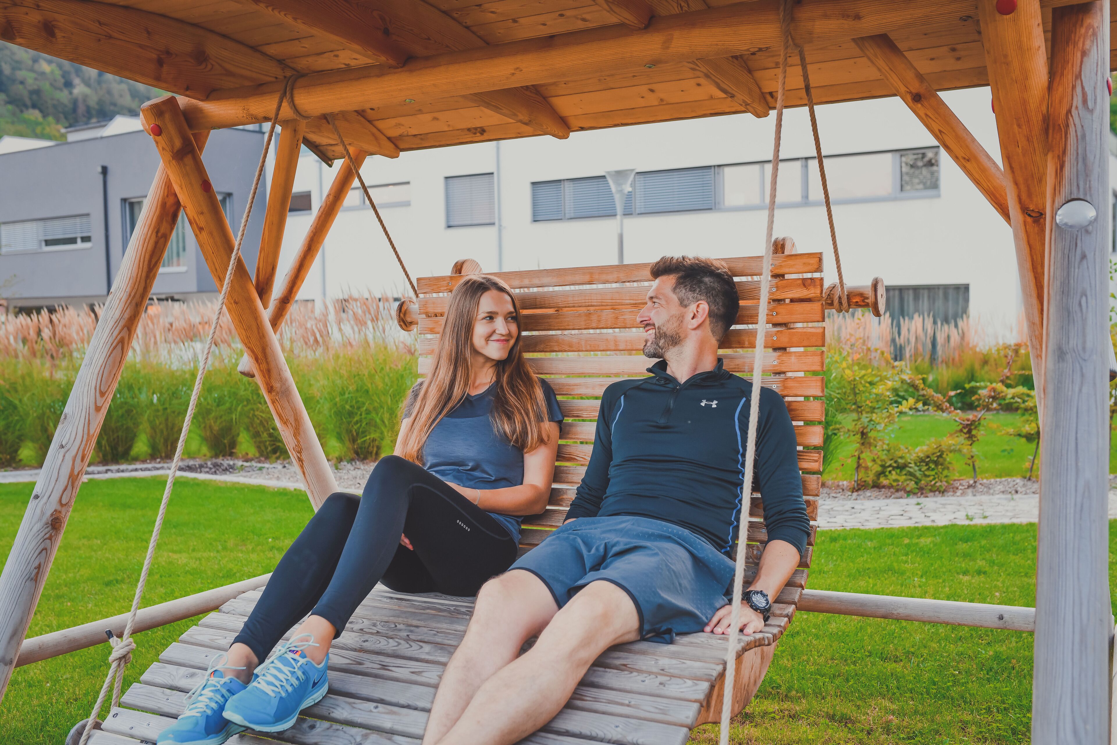 A wooden swing bench with a metal frame, set in a garden with grass and plants.