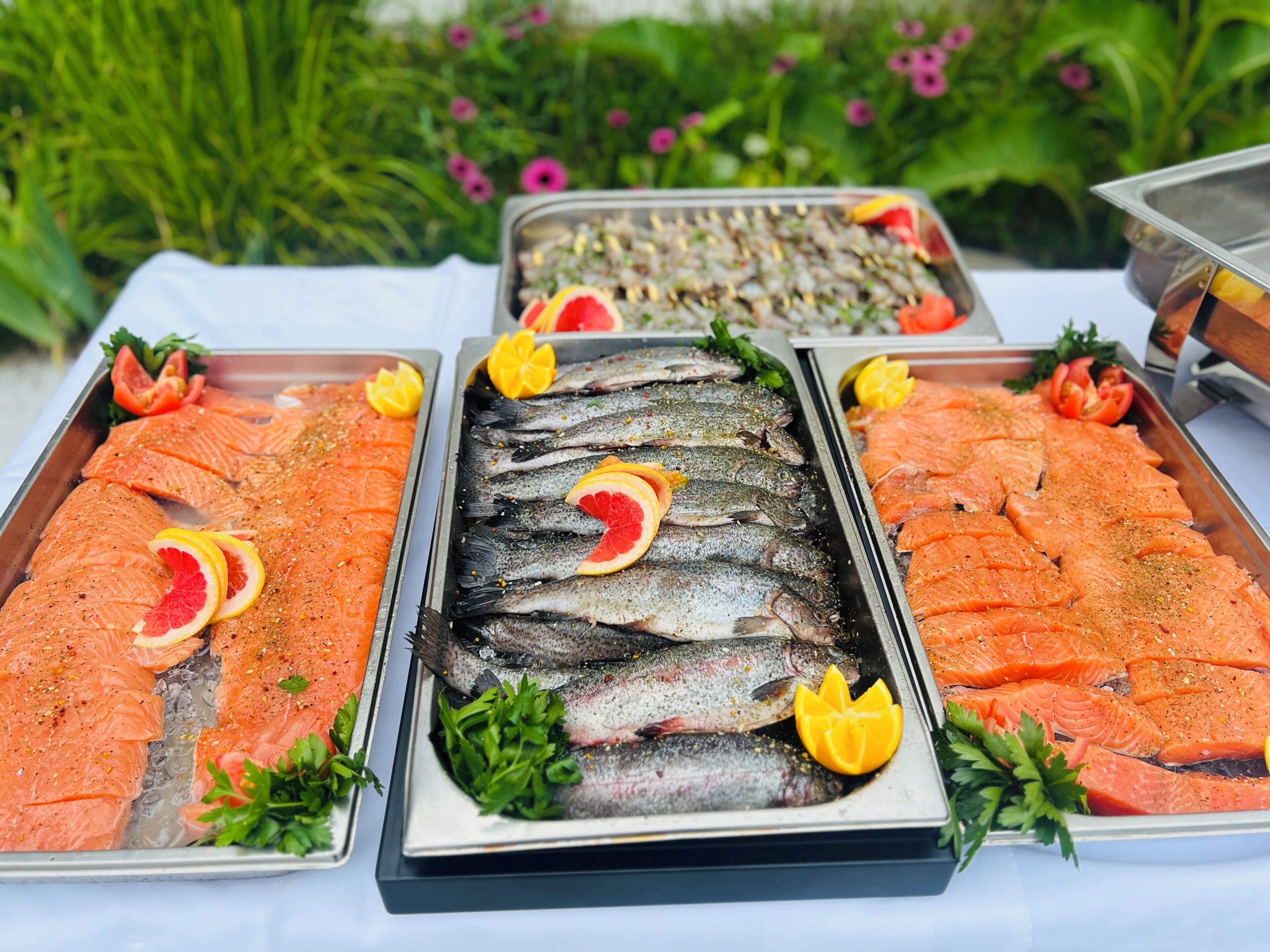 Drei Tabletts mit Lachs und Forelle auf einem Tisch, im Hintergrund ein Garten.
