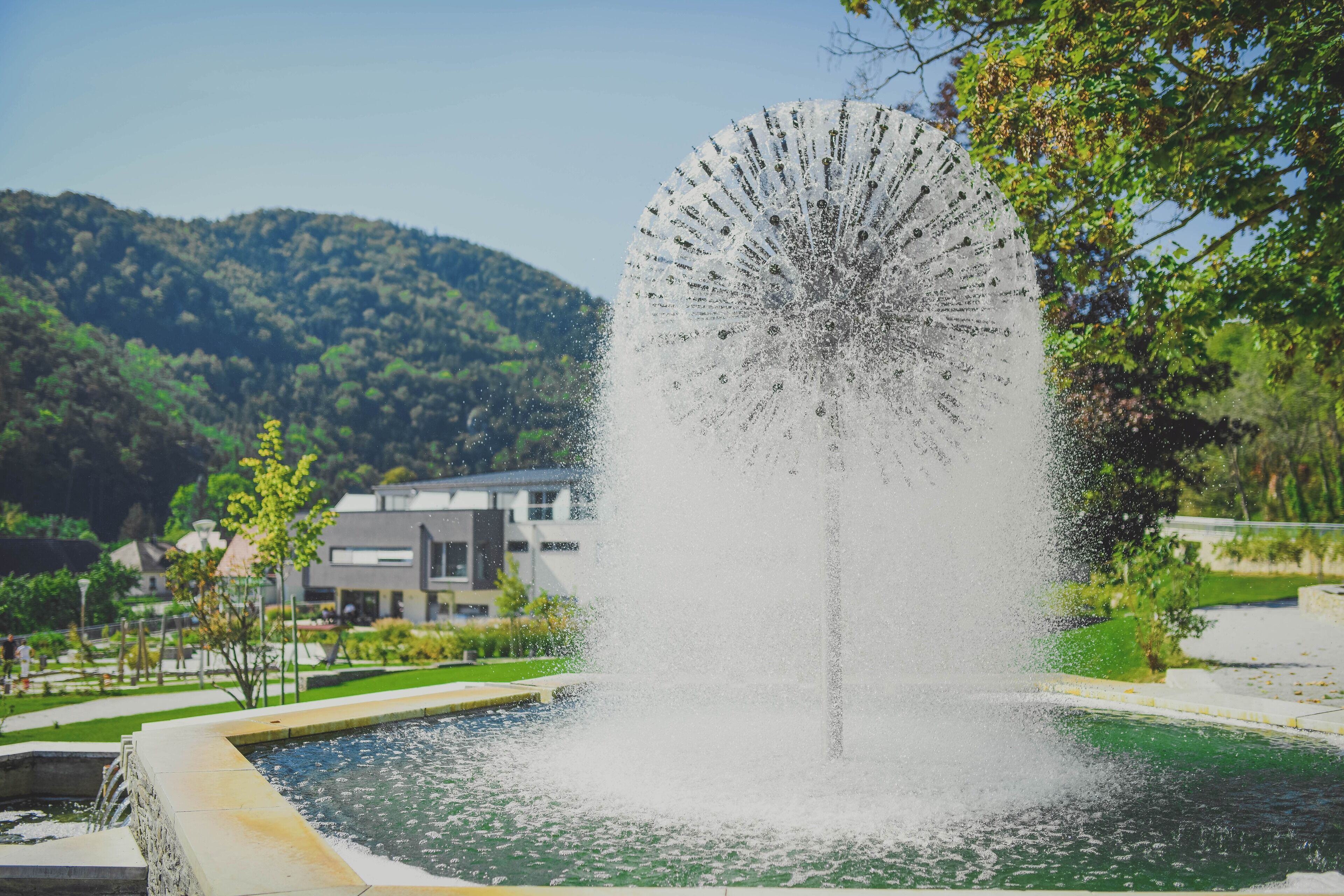 A fountain with water jets in a landscaped area with a pool, surrounded by greenery and buildings.