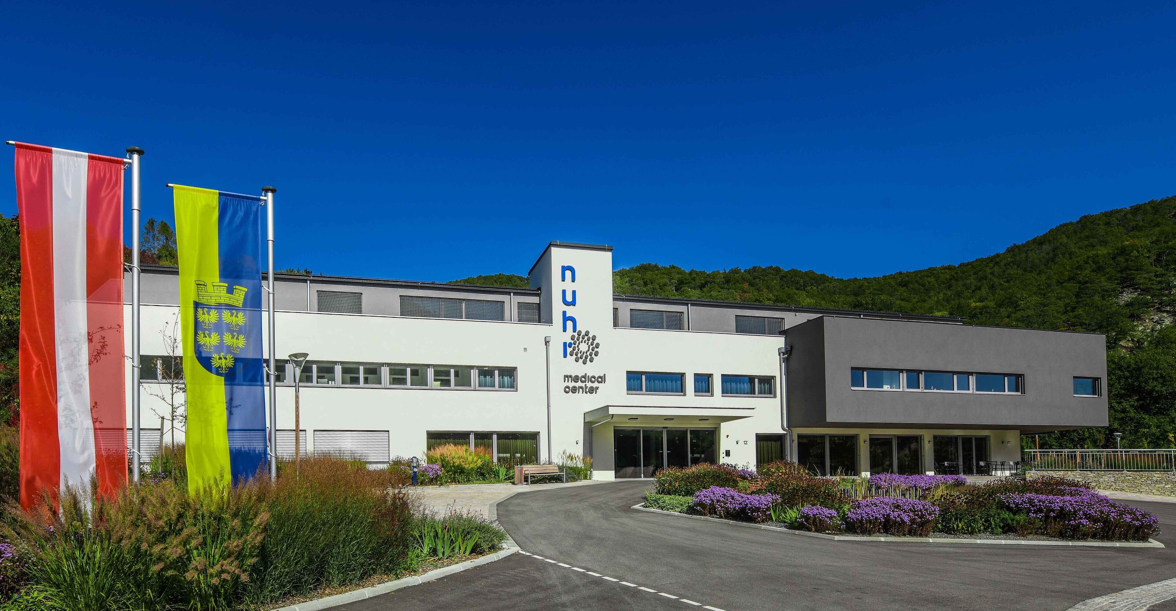 A medical center with a modern building, a parking area, and a landscaped garden with flowers.