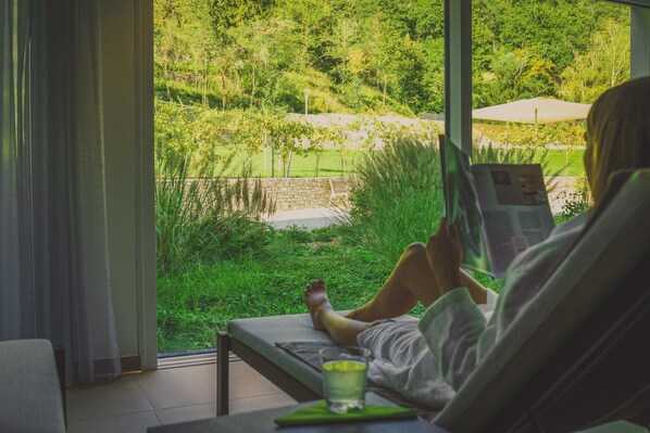A person sitting on a bed by a window with a view of a garden. - NUHR Medical - Hotel & Restaurant (Senftenberg)