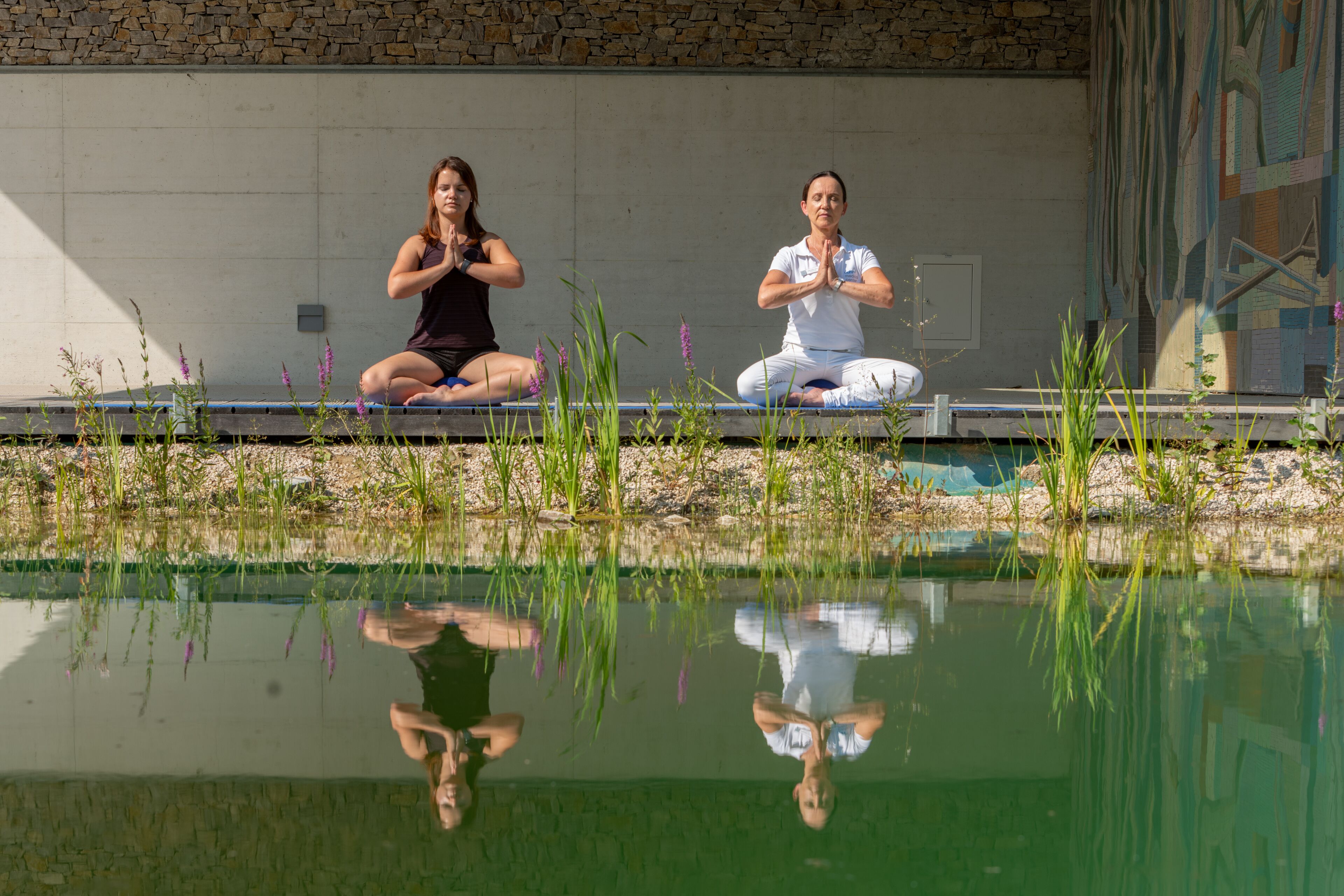 Two individuals seated on a platform by a reflective water body, with a stone wall and a mural in the background.