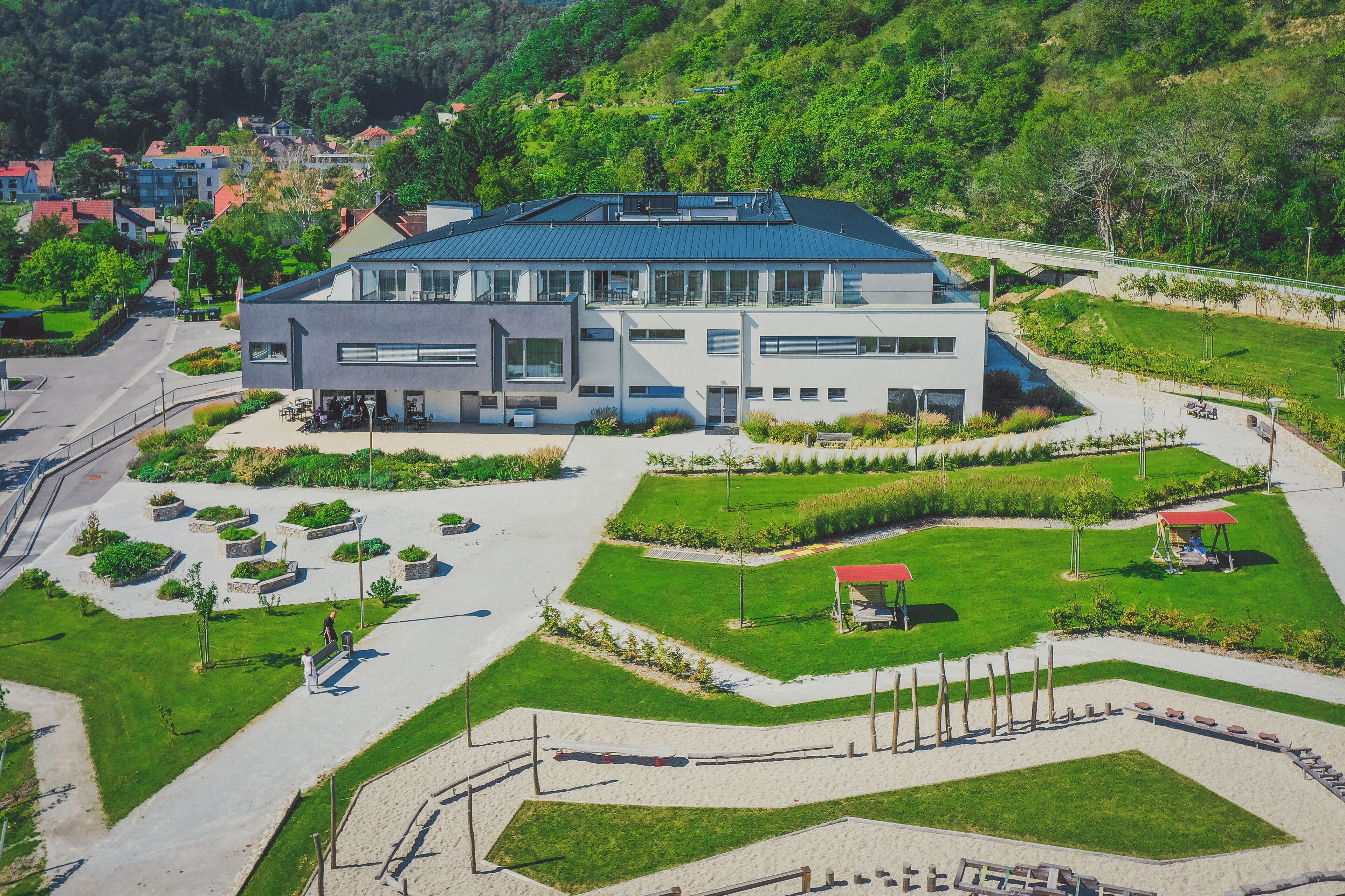 Een modern gebouw met een plat dak, omringd door een aangelegde tuin met paden, banken en een speeltuintje.