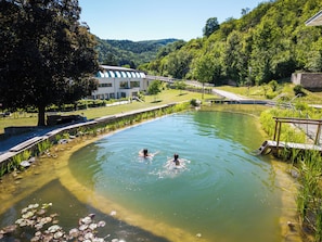 Piscina com duas pessoas, rodeada por vegetação e um edifício.