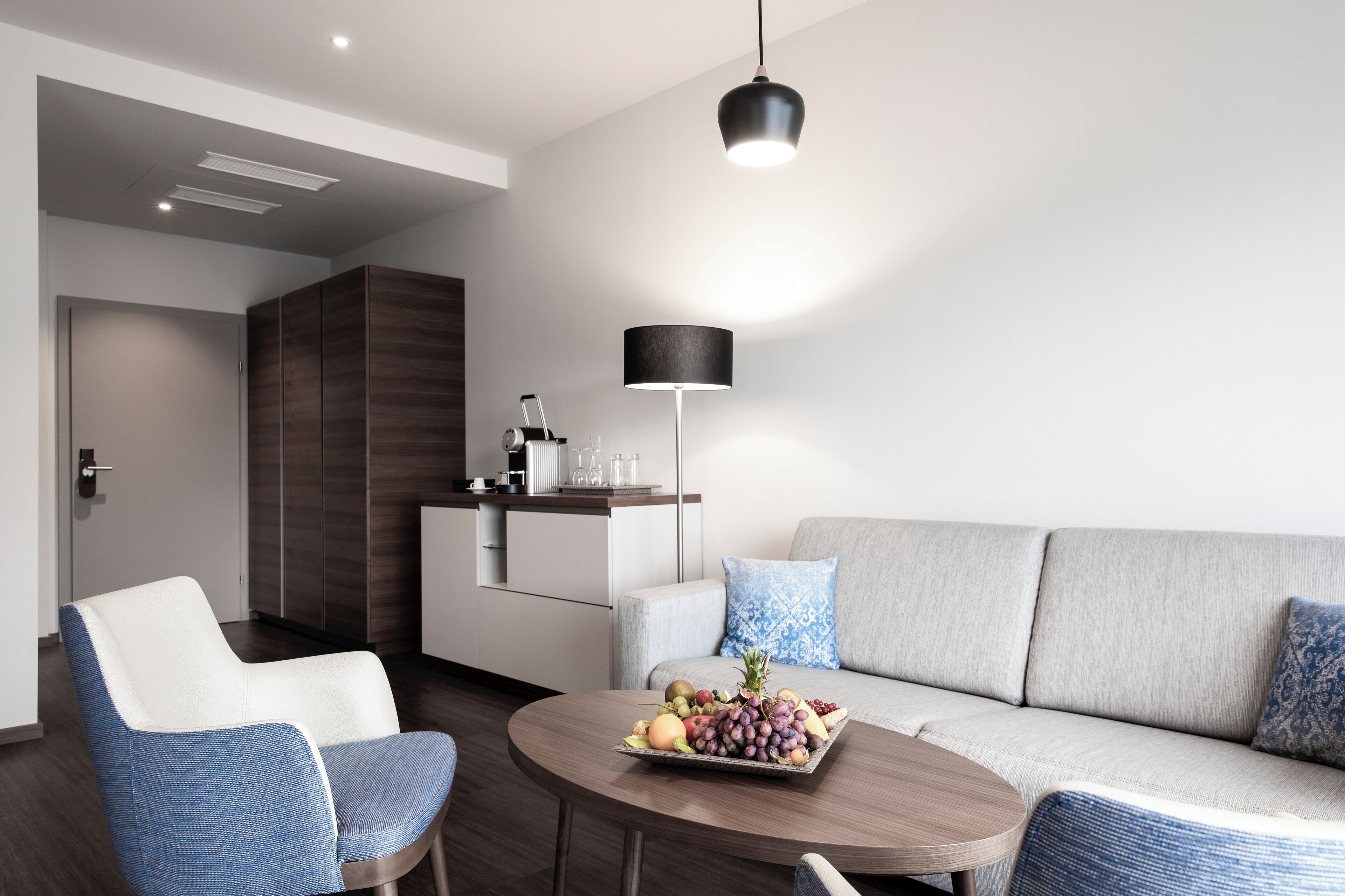 Un salon moderne avec un canapé, une table basse sur laquelle se trouvent des fruits, un fauteuil, une lampe et, au fond, un coin cuisine.