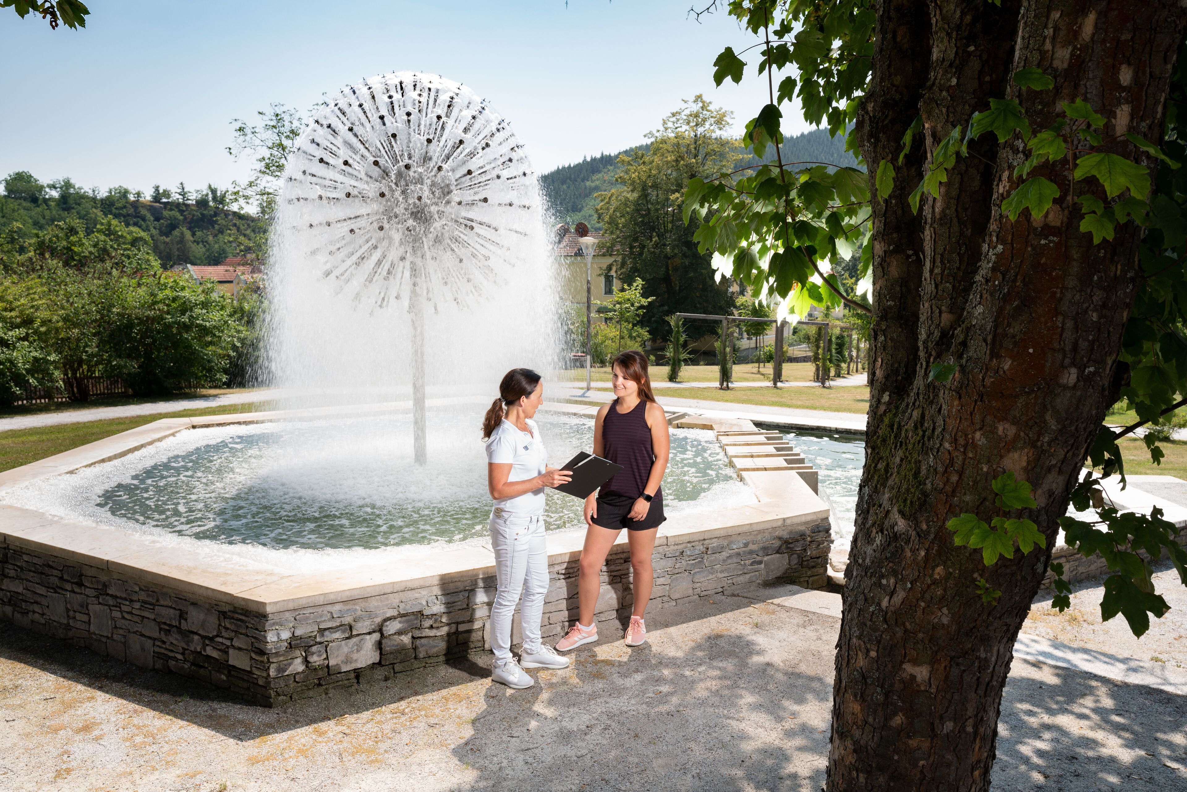 Two people standing by a fountain with a water jet in a park.