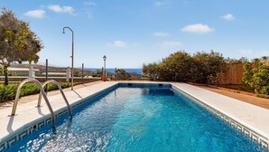 A rectangular swimming pool with a metal ladder on one side, surrounded by a concrete deck and a low stone wall.