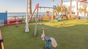 A playground with swings, slides, and a climbing structure near a white building with a view of the sea.