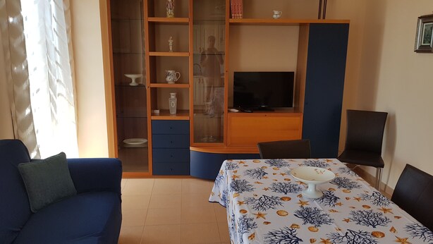 Ein Zimmer mit einem blauen Sofa, einem Esstisch mit Tischdecke, einem Fernseher auf einem Schrank, einem Schrank mit Glastüren und einem Bücherregal.