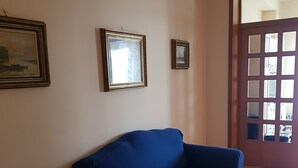 Ein Zimmer mit einem blauen Sofa, zwei gerahmten Bildern an der Wand und einem Fenster mit Blick ins Freie.