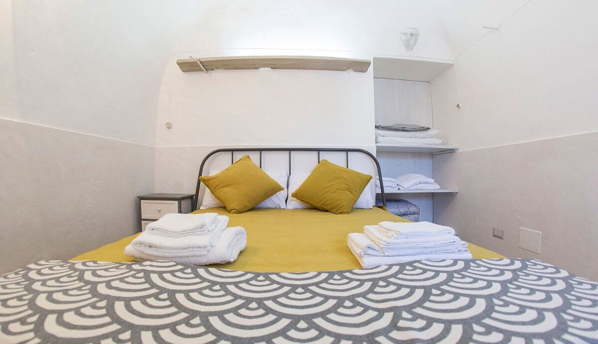 A hotel room with a bed, two pillows, a bedside table, a shelf with folded towels, and a patterned bedspread.