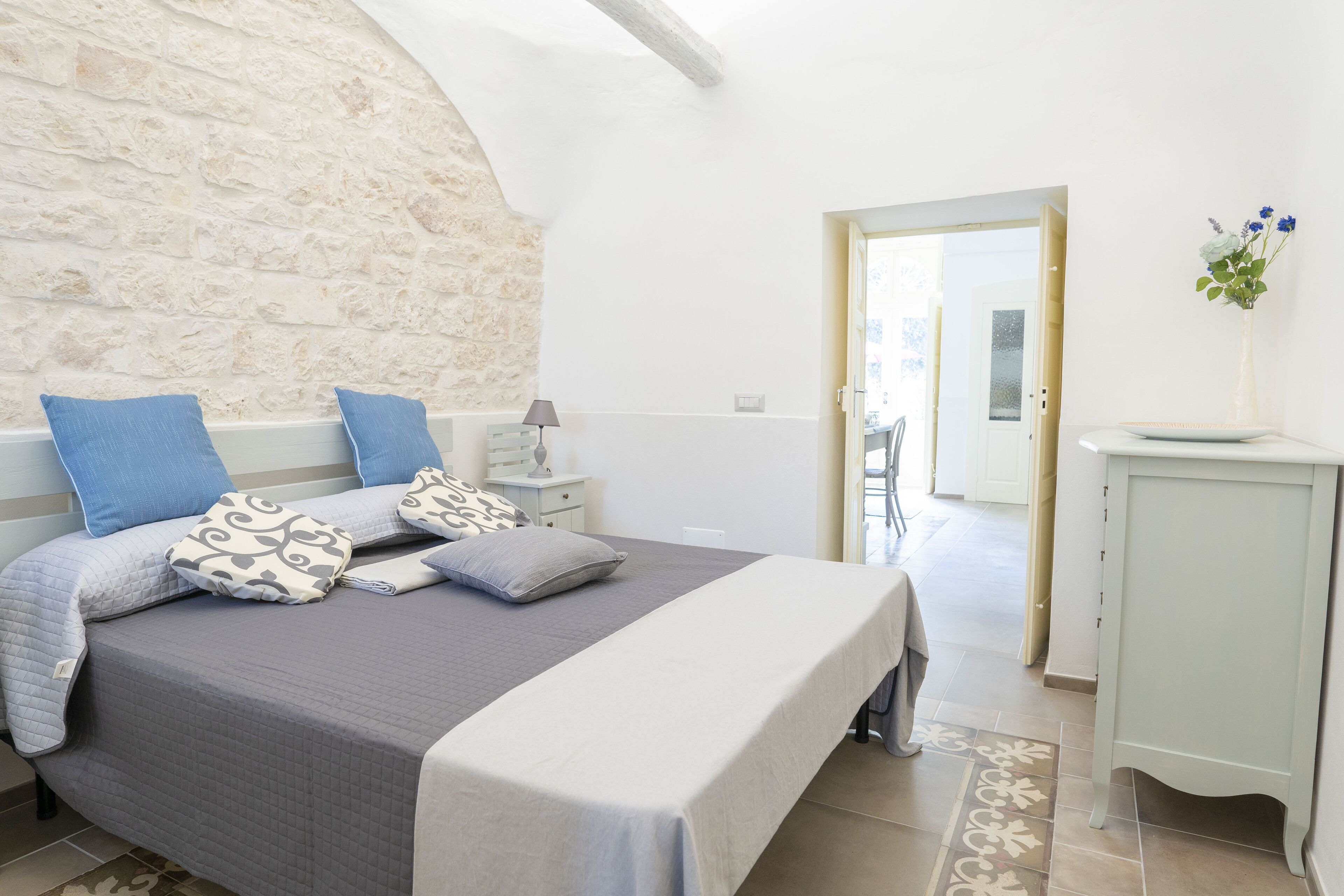 Un dormitorio con pared de piedra, una cama con almohadas estampadas, una mesita de noche con una lámpara y una puerta que da a otra habitación.